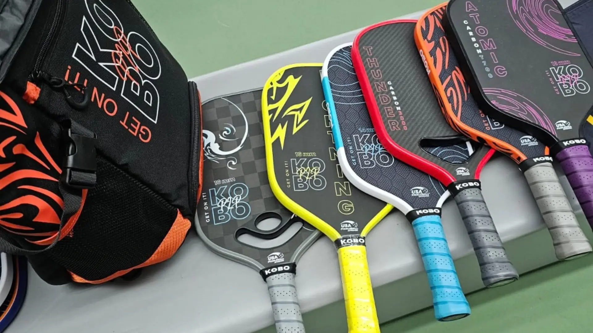 Selection of KOBO pickleball paddles arranged beside a KOBO paddle bag on a pickleball court